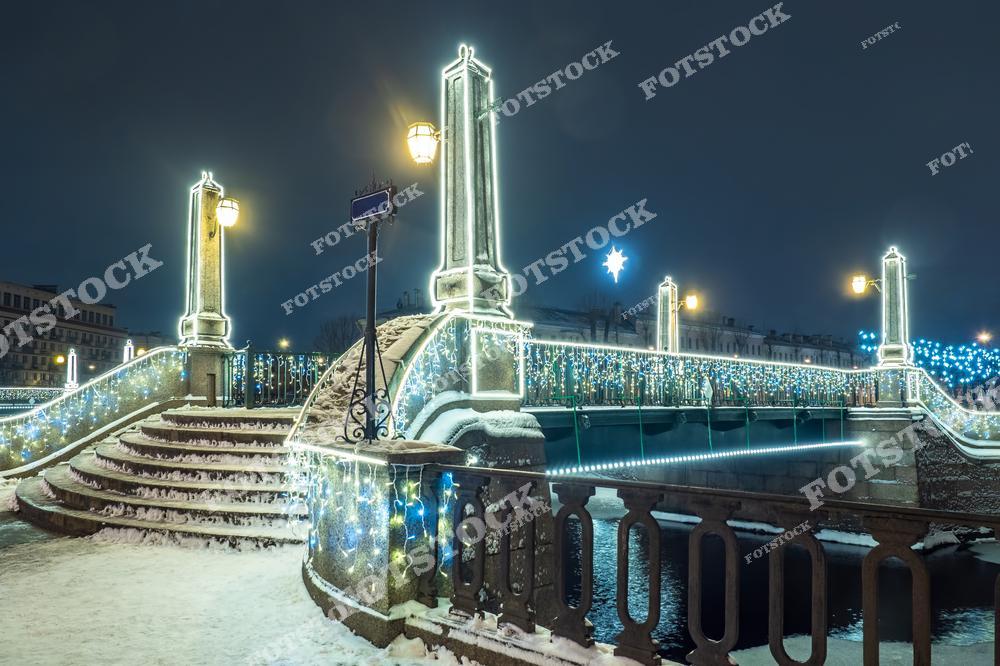На фотографии изображен Красногвардейский мост через канал Грибоедова в Санкт-Петербурге, украшенный к Новому году. Светящиеся гирлянды огибают перила и столбы моста, придавая ему праздничный вид. Легкий слой снега покрывает ступени и дорожки вокруг, а огни фонарей добавляют теплый свет в зимний пейзаж. Мост, расположенный в центре города, становится ярким элементом праздничной атмосферы Санкт-Петербурга.